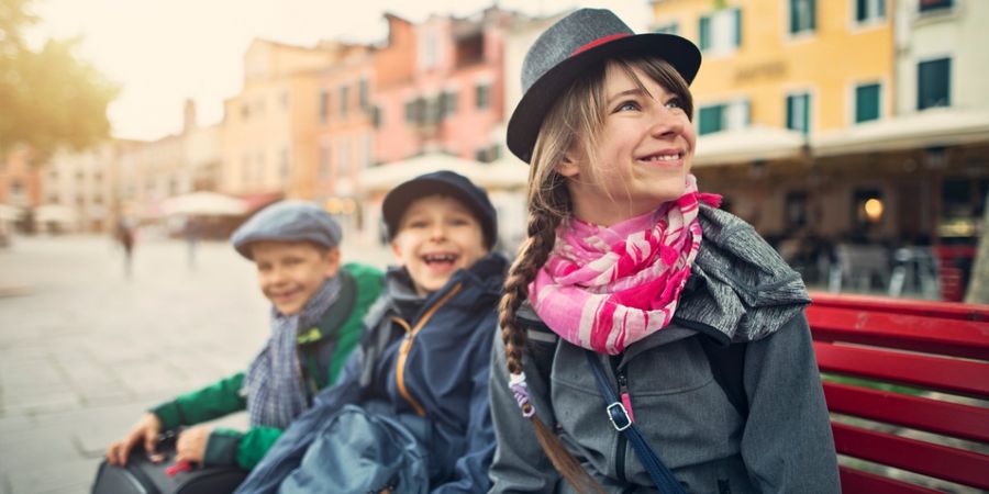 Een foto van drie kinderen die op een bankje voor felgekleurde gebouwen zitten.