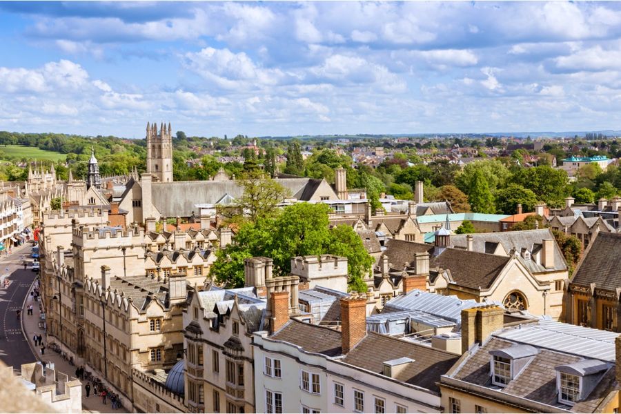 Een foto van de daken van Oxford en de klokkentoren van de kathedraal van de stad, met in de verte bomen en heuvels.