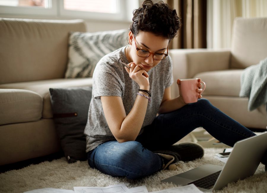 Een vrouw op de grond, voor haar laptop, met een pen in haar ene hand en een kopje koffie in de andere.