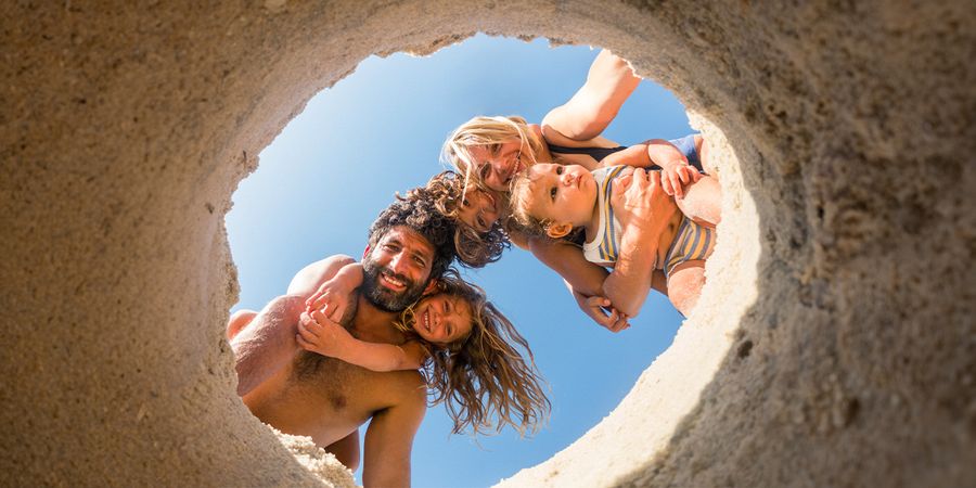 Een gezin van vier op het strand – mama, papa, dochter en babyzoon – kijkt in een gat dat in het zand is gegraven, een speels zomermoment dat inspireert voor strandfoto’s en strandfotografie.