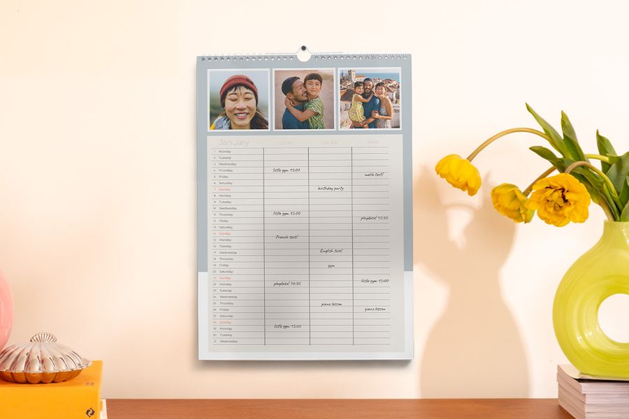 Een familieplanner hangt aan de muur naast een vaas en enkele boeken, ter illustratie van albelli's tip om het schooljaar georganiseerd te starten met een persoonlijke planner.