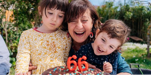 Een vrouw glimlacht naast haar verjaardagstaart met kaarsjes die '66' aangeven, geflankeerd door een jong meisje en een jonge jongen, ter introductie van albelli's ultieme cadeaugids voor verjaardagen.