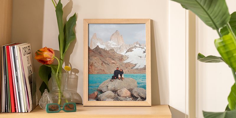 Een ingelijste landschapsfoto op een houten plank naast boeken en bloemen in een vaas, als voorbeeld van een schilderij ophangen zonder boren bij albelli.