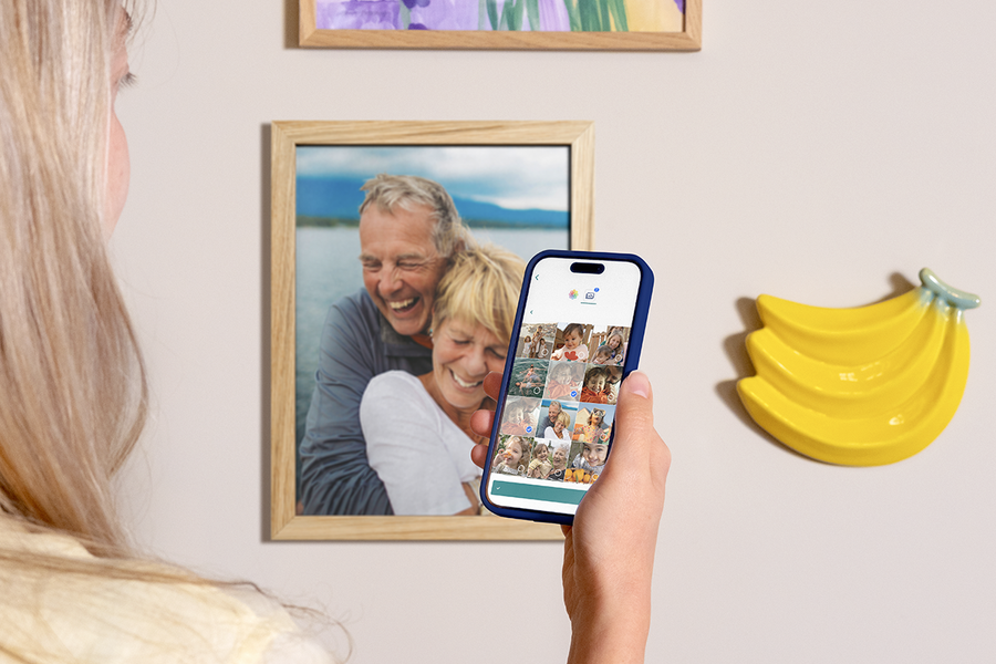 Een vrouw houdt haar telefoon omhoog om te laten zien hoe makkelijk het is om albelli foto afdrukken te bestellen via de app, met ingelijste foto afdrukken aan de muur achter haar, als inspiratie voor online foto afdrukken en foto’s afdrukken vanaf je telefoon.
