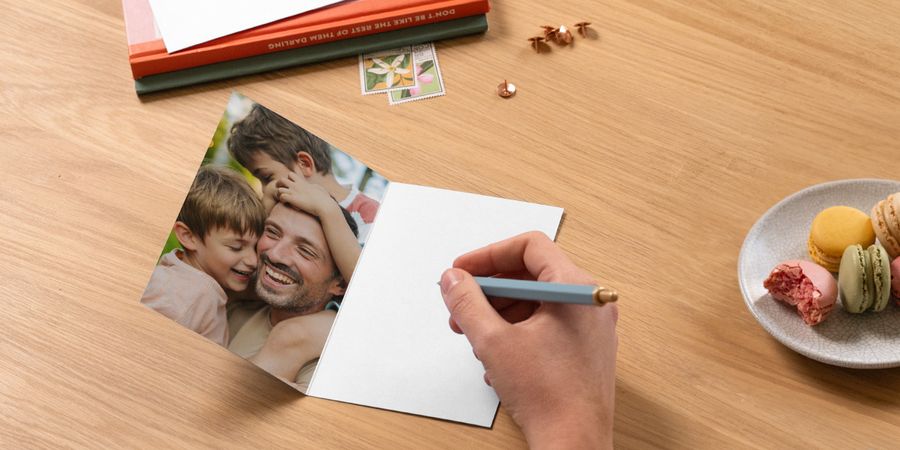 Een open gepersonaliseerde albelli vaderdagkaart toont een vader met zijn twee zoons, met een pen klaar om een boodschap te schrijven naast boeken en een bord macarons, als inspiratie voor wat je op een vaderdagkaart schrijft.