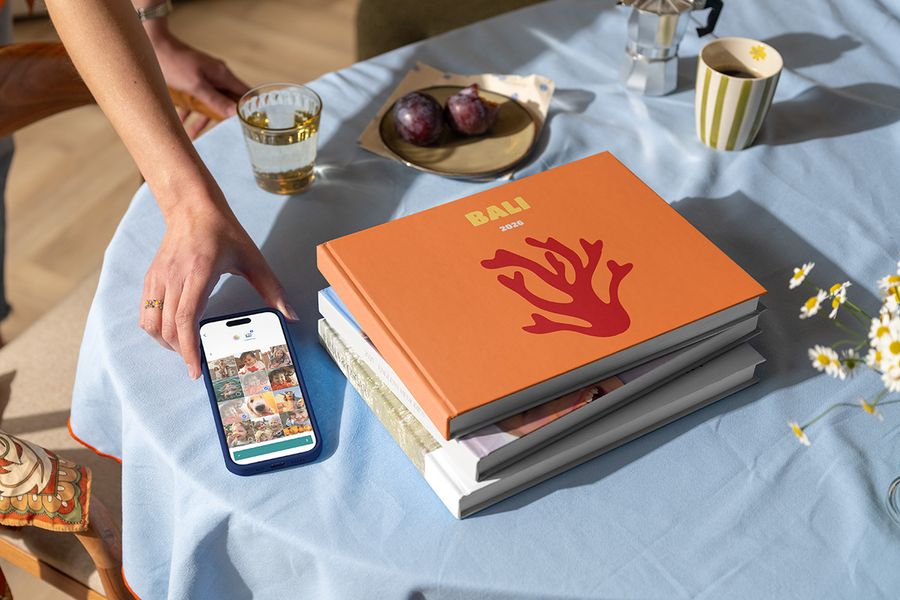 Een stapel verschillende albelli reisfotoboeken staat naast een hand die een telefoon vasthoudt met een vakantiefoto klaar om toe te voegen, met bovenop een vakantie fotoboek Bali 2026 met koraal op de cover, als illustratie van hoe je een gepersonaliseerd vakantie fotoboek maakt.