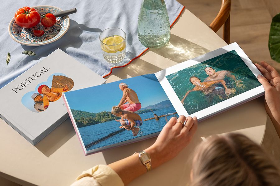 Twee verschillende albelli reisfotoboeken zijn te zien: een vrouw bladert door een open vakantie fotoboek met familiefoto’s aan zee, naast een gesloten Portugal vakantiealbum, als illustratie van een gepersonaliseerd vakantie fotoalbum vol reisherinneringen.