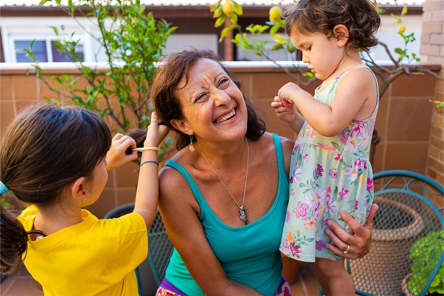 Een vrouw zit op een balkon terwijl twee jonge meisjes met haar haar spelen en het stylen, een vrolijk moment uit albelli's tips voor een vakantie thuis met kinderen.