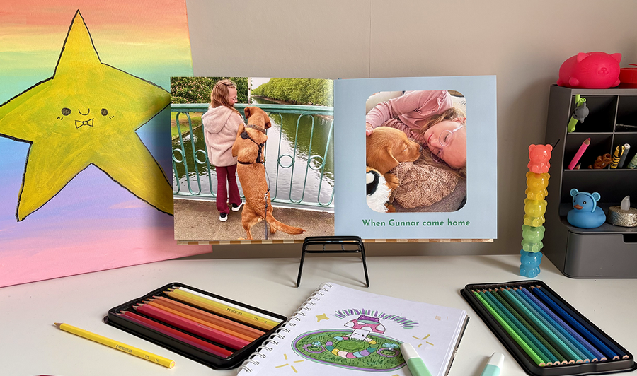 Een geopend kinderfotoboek ligt op het bureau van een kind, omringd door kleurpotloden en kunstwerken, met op de linkerpagina een foto van een meisje met haar hond en op de rechterpagina nog een afbeelding met bijschrift, ter illustratie van albelli's tips voor het combineren van foto's en kunstwerken in een familiefotoboek.