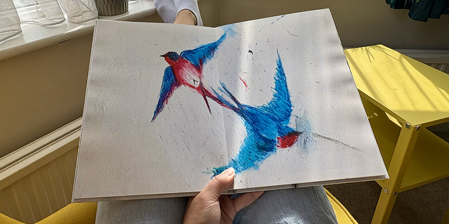Een geopend kinderfotoboek met een volledige paginaspreiding van een vogeltekening ligt op de schoot van een vrouw terwijl ze in een fauteuil zit, ter illustratie van albelli's stappen voor het maken van een familiefotoboek met de tekeningen van je kind.