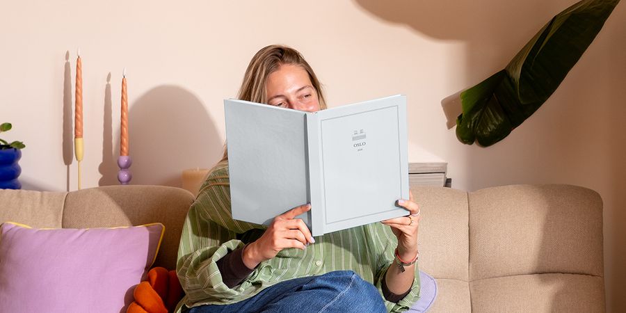 Een vrouw zit op de bank naast een kleurrijk kussen en bekijkt een gepersonaliseerd reisfotoboek met de titel 'Oslo', ter illustratie van albelli's nieuwe Summer Travel Series ontwerpen.