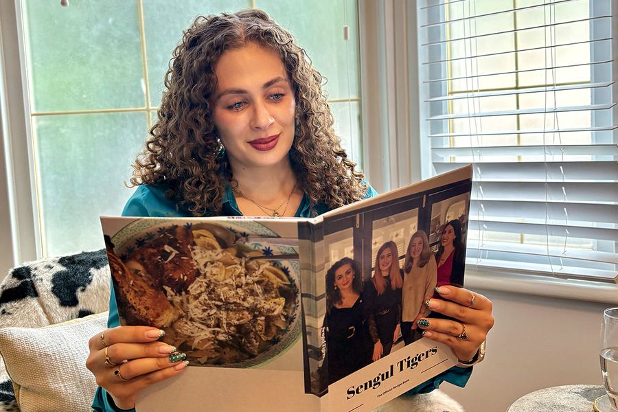 Sarah, die bij albelli werkt, zit in een koeienprint stoel en bekijkt haar gepersonaliseerde familiekookboek, waarvan de cover vier vrouwen toont, waaronder Sarah zelf, ter illustratie van hoe een fotoboek met recepten familierecepten en herinneringen samenbrengt.