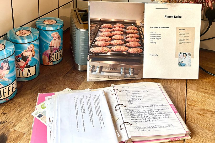 Sarah's geopende gepersonaliseerde kookboek toont een recept voor haar Nene's kofte, geplaatst voor een oude map met handgeschreven familierecepten, beide liggend op een keukenblad, ter illustratie van hoe zij generaties recepten in één boek samenbracht.
