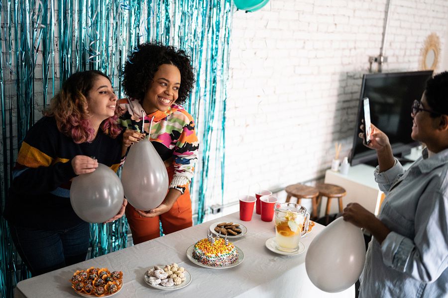 Twee meisjes poseren voor een glitterfoto-achtergrond, met ballonnen in de hand bij een snacktafel vol feestvoedsel en een verjaardagstaart, ter illustratie van albelli's tips om de feestdecoratie en het buffet te fotograferen.