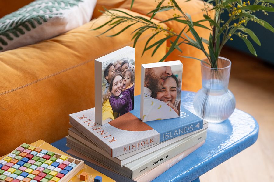 Twee gepersonaliseerde fotoblokken rusten op een selectie koffietafelboeken op een blauw bijzettafeltje naast een oranje fluwelen bank, ter illustratie van hoe albelli fotoblokken een persoonlijke toets geven aan een hygge-huis.