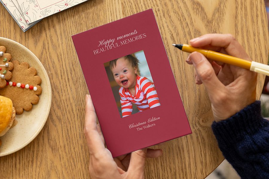 Handen die klaarstaan om een gepersonaliseerde fotokaart te ondertekenen met een lachende baby, een kerstboodschap en een familienaam op de voorkant, naast een bord kerstkoekjes op een tafel, ter illustratie van albelli’s ideeën voor gepersonaliseerde kerstkaarten voor de eerste kerst van een baby.