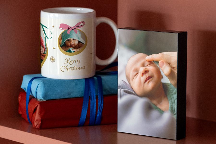 Gepersonaliseerde mok met een foto van een baby in een handdoek en een kerstwens, op twee ingepakte cadeaus naast een fotoblok van een slapende pasgeborene, ter illustratie van albelli’s ideeën voor fotocadeaus voor de eerste kerst van jonge ouders.