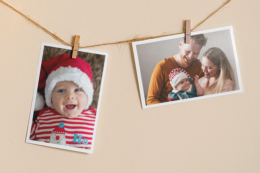 Fotoprints die met wasknijpers aan een touw hangen, waarvan er één een lachende baby met een kerstmuts toont en de andere een lachende man en vrouw die een baby in een kerstoutfit vasthouden, ter illustratie van albelli’s tips om van een favoriete foto een zelfgemaakte kersthanger te maken.