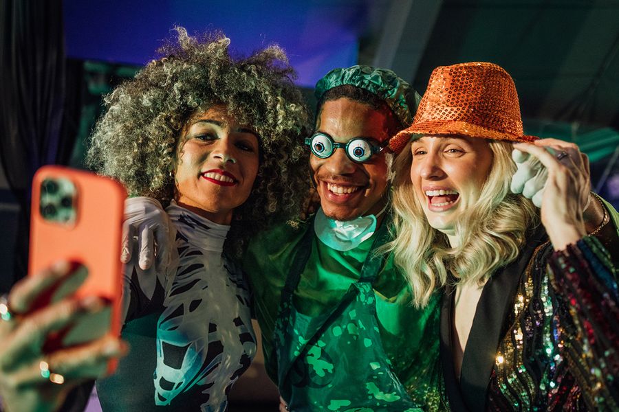 Drie volwassenen poseren voor een selfie op een Halloweenfeest, een vrouw in een skeletkostuum, een man verkleed als enge chirurg met een grappige bril en een vrouw met een hoed vol pailletten, ter illustratie van albelli's Halloween-teksten voor groepen.