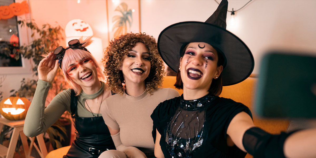 Drie vrouwen in verkleedkleding op een Halloweenfeest, een heks, een dier en een cyberpunk, poseren voor een selfie op hun telefoon, ter introductie van albelli's 105 Halloween-teksten voor Instagram.
