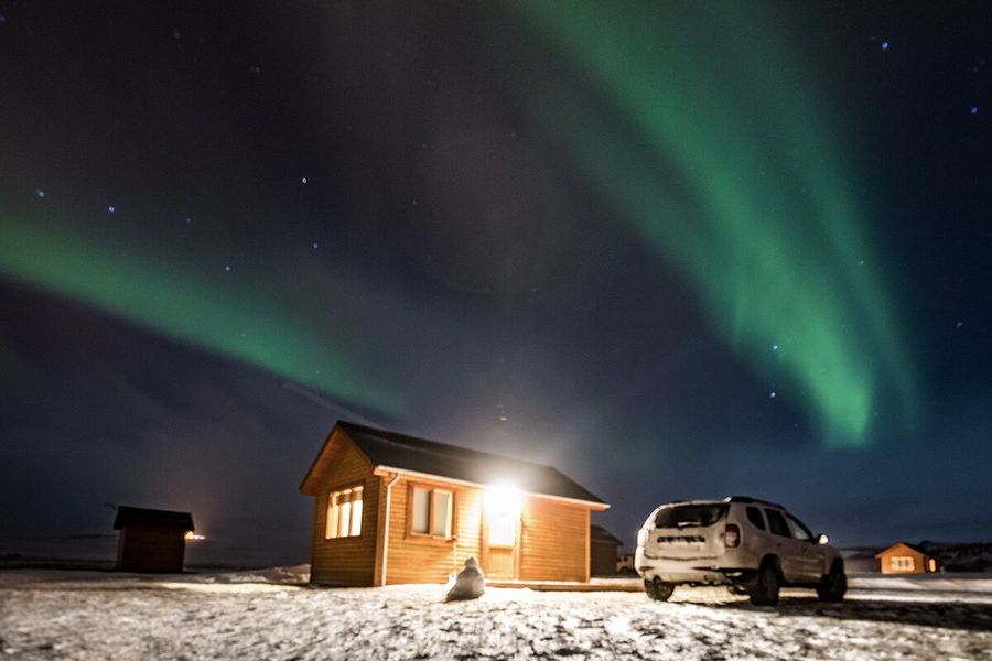 Een hut met licht dat naar buiten schijnt, naast een auto in een zeer besneeuwd landschap, met groen noorderlicht aan de hemel erboven, ter illustratie van albelli’s uitleg over wat het noorderlicht is en wat aurora borealis veroorzaakt.