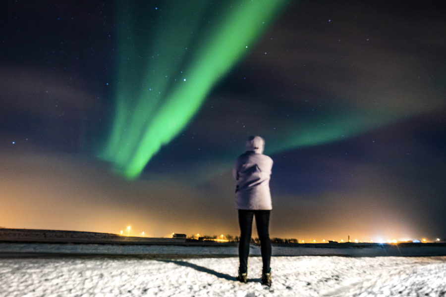 Een persoon die staat in een besneeuwd landschap en naar het groene noorderlicht aan de hemel kijkt, ter illustratie van albelli’s tips om noorderlicht met een smartphone te fotograferen.
