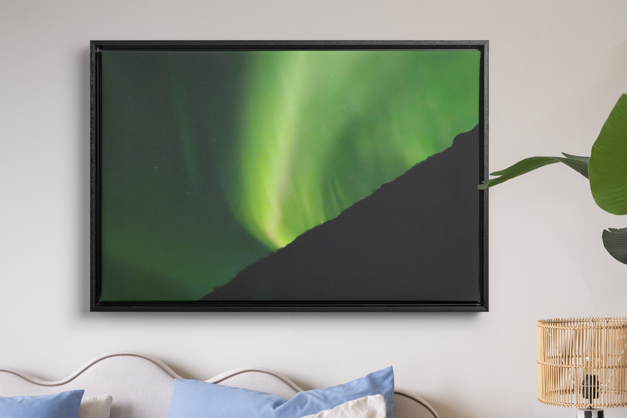 Een ingelijste print van zeer groen noorderlicht hangt aan de muur boven een bed en een raffialamp, ter illustratie van albelli’s wanddecoratie om noorderlichtfoto’s om te zetten in iets om te bewaren.