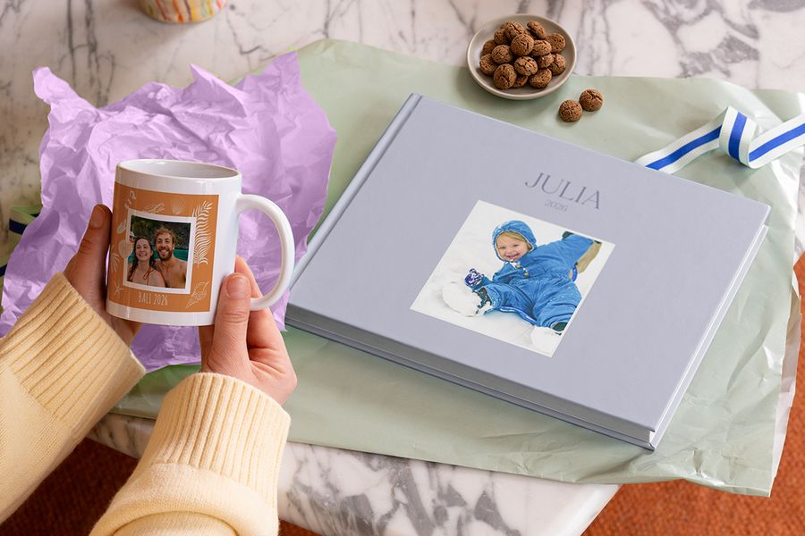 Een hand die een mok met ‘BALI 2026’ vasthoudt naast een grijsblauw hardcover fotoboek met ‘JULIA 2026’ op de kaft, met een jong kind in een sneeuwpak op de foto en pepernoten op een marmeren tafel, ter illustratie van albelli’s uitleg waarom een gepersonaliseerd Sinterklaascadeau jaren plezier geeft.