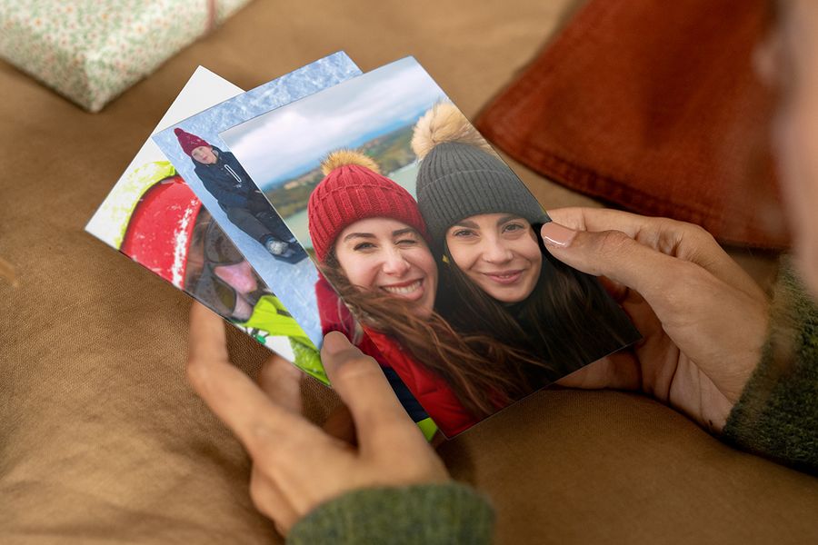 Persoon die fotoprints in de handen houdt, waarbij op de voorste print twee lachende vrouwen met wintermutsen en winterjassen te zien zijn, ter illustratie van albelli’s tips om de juiste foto’s te vinden voor een gepersonaliseerd fotocadeau.