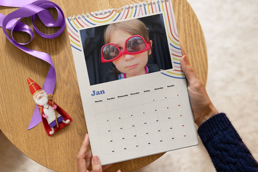 Een jong kind met een grote rode zonnebril op de foto bovenaan een kleurrijke wandkalender voor januari, met een paars lint en een klein Sinterklaasfiguurtje ernaast, ter illustratie van albelli’s tip voor een persoonlijke fotokalender als Sinterklaascadeau.