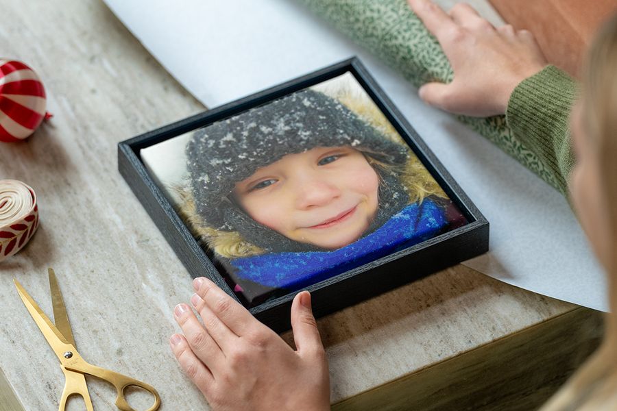 Ingelijst canvas van een jong meisje in winterkleding in de sneeuw, met de handen van een vrouw die klaar zijn om het in te pakken, naast inpakpapier, een schaar en lint, ter illustratie van albelli’s tips om een gepersonaliseerd fotocadeau te kiezen dat past bij de stijl van de ontvanger.