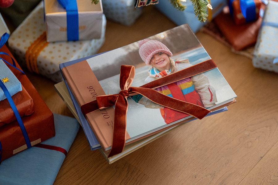 Een stapel fotoboeken met een fluwelen lint eromheen, waarvan het bovenste boek een lachend meisje in een roze gebreide muts toont en ‘2026’ op de rug, tussen ingepakte kerstcadeaus, ter illustratie van albelli’s fotoboek van het jaar.